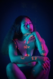 A woman sits in a contemplative pose, with her hand resting on her chin. She is illuminated by vibrant lighting that casts shades of blue, teal, and pink on her skin. Chinese characters are projected onto her body, adding an intriguing and artistic element to the scene. Her expression is pensive, and the overall composition combines elements of mystery and introspection.
