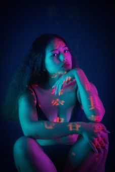 A woman sits in a contemplative pose, with her hand resting on her chin. She is illuminated by vibrant lighting that casts shades of blue, teal, and pink on her skin. Chinese characters are projected onto her body, adding an intriguing and artistic element to the scene. Her expression is pensive, and the overall composition combines elements of mystery and introspection.