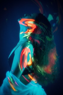 A person is posed with their head tilted back, enveloped in an array of colorful light projections. The vivid swirls of neon colors, including red, orange, green, and blue, create an abstract pattern across their skin and hair, forming a captivating visual effect against a dark background. The individual appears to be wearing a dark garment with a light fabric draped around them.