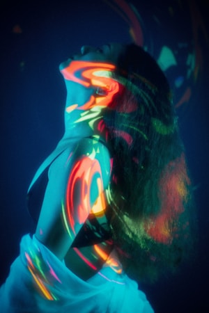 A person is posed with their head tilted back, enveloped in an array of colorful light projections. The vivid swirls of neon colors, including red, orange, green, and blue, create an abstract pattern across their skin and hair, forming a captivating visual effect against a dark background. The individual appears to be wearing a dark garment with a light fabric draped around them.