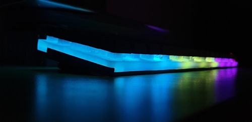 A close-up side view of a mechanical keyboard with RGB backlighting, displaying a spectrum of colors including blue, green, and purple. The lighting casts a vivid glow on the keyboard's surface, creating a dramatic contrast against the dark background.