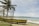 A yellow boat with red lettering sits on a sandy beach next to a wire fence. Palm trees sway gently in the background, with the ocean stretching to the horizon under a partly cloudy sky.