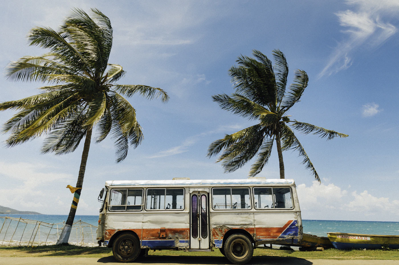 A vintage Jamaican minibus parked between two palm trees on a beachfront, with a small Jamaican-flag ribbon tied to a palm trunk