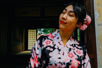 A woman wearing a traditional floral-patterned garment is leaning against a wooden doorframe, with her eyes closed and a peaceful expression. Her hair is adorned with pink flowers, and the background features a dimly lit, traditional interior with paper windows.