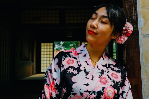 A woman wearing a traditional floral-patterned garment is leaning against a wooden doorframe, with her eyes closed and a peaceful expression. Her hair is adorned with pink flowers, and the background features a dimly lit, traditional interior with paper windows.