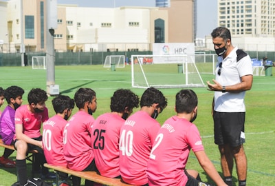 A soccer coach engages players seated on a bench during what seems like a break or team talk on a green soccer field. The players wear matching pink jerseys with numbers, while the coach is dressed in a white polo shirt. Small soccer goals and buildings appear in the background.