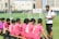 A soccer coach engages players seated on a bench during what seems like a break or team talk on a green soccer field. The players wear matching pink jerseys with numbers, while the coach is dressed in a white polo shirt. Small soccer goals and buildings appear in the background.
