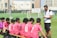 A soccer coach engages players seated on a bench during what seems like a break or team talk on a green soccer field. The players wear matching pink jerseys with numbers, while the coach is dressed in a white polo shirt. Small soccer goals and buildings appear in the background.