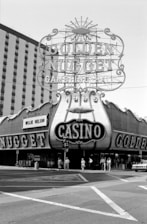A large vintage neon sign advertises the Golden Nugget gambling hall and casino, with ornate lettering and decorative elements. The street scene features several people walking past the entrance, and a car is parked nearby. A high-rise building is visible in the background.