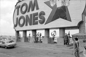 A large sign with the name 'Tom Jones' in bold letters is displayed above a group of people outside. The platform below the sign has several individuals dressed in Roman-inspired attire, holding spears and shields. A classic car is parked to the left, and a sign indicating 'Self Parking Entrance' is on the right. The background shows a vast parking lot filled with cars under an overcast sky.
