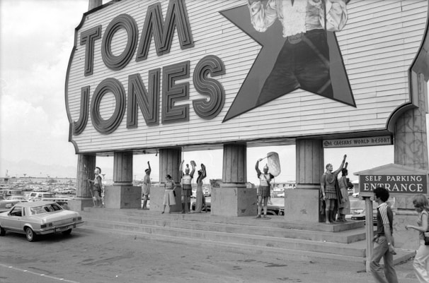 A large sign with the name 'Tom Jones' in bold letters is displayed above a group of people outside. The platform below the sign has several individuals dressed in Roman-inspired attire, holding spears and shields. A classic car is parked to the left, and a sign indicating 'Self Parking Entrance' is on the right. The background shows a vast parking lot filled with cars under an overcast sky.