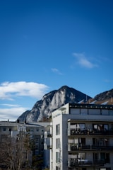 A modern apartment building stands in the foreground with multiple balconies. Behind it, a large mountain with rugged, rocky features rises against a clear blue sky. A few trees, mostly barren, are visible near the buildings.