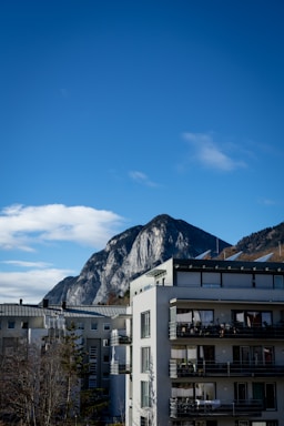 A modern apartment building stands in the foreground with multiple balconies. Behind it, a large mountain with rugged, rocky features rises against a clear blue sky. A few trees, mostly barren, are visible near the buildings.