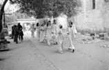 A group of people dressed in formal attire, possibly for a wedding, walk along a stone pathway. Bridesmaids wear matching gowns and hold bouquets, while groomsmen wear white tuxedos with bowties. The scene is outdoors, framed by trees and ancient stone buildings, with a background of other casually dressed people walking in the opposite direction.