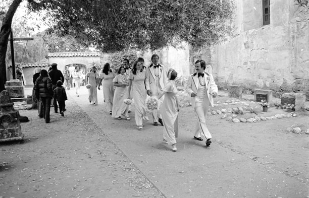 A group of people dressed in formal attire, possibly for a wedding, walk along a stone pathway. Bridesmaids wear matching gowns and hold bouquets, while groomsmen wear white tuxedos with bowties. The scene is outdoors, framed by trees and ancient stone buildings, with a background of other casually dressed people walking in the opposite direction.