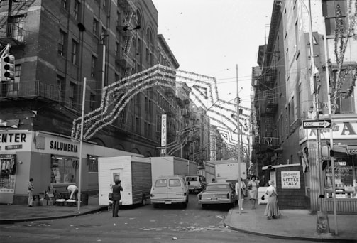 A black and white photograph of a busy street scene in Little Italy with buildings decorated with hanging festive garlands. Several vehicles, including vans and cars, line the street, and pedestrians can be seen walking along the sidewalks. Storefronts with various signs and fire escapes on the buildings add to the urban atmosphere.