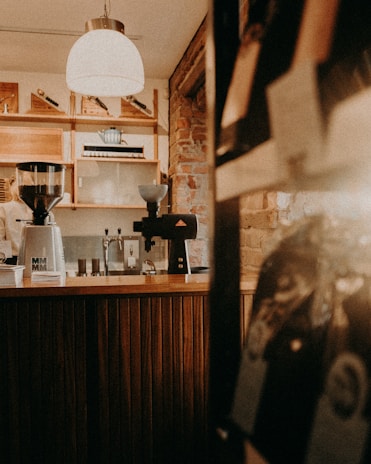 A cozy coffee shop interior with wooden paneling and exposed brick walls. A large pendant light hangs from the ceiling, illuminating the counter area. Various coffee-making equipment is visible, including a coffee grinder with a container labeled 'MMM' and an espresso machine. Shelves with decorative items and cups are in the background.