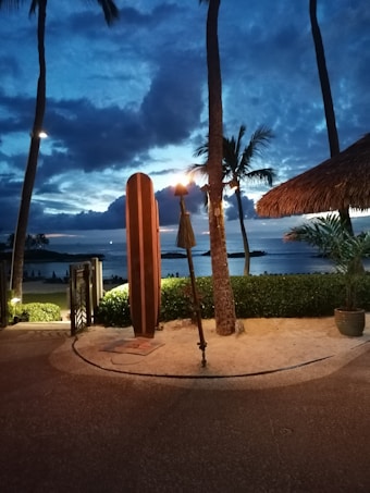 A tranquil tropical beach scene at dusk features a tall tiki torch next to a surfboard statue, surrounded by palm trees. The sky is a mix of deep blues and soft yellows as the sun sets over the ocean, with lush greenery framing the foreground.