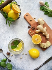 A selection of fresh ingredients including lemons, ginger, and mint is arranged artistically on a textured surface. A glass of water garnished with a lemon slice and mint leaves is placed nearby, while a wooden cutting board holds slices of ginger and a halved lemon. The scene is complemented by a honey dipper and some star anise, creating a refreshing and natural aesthetic.