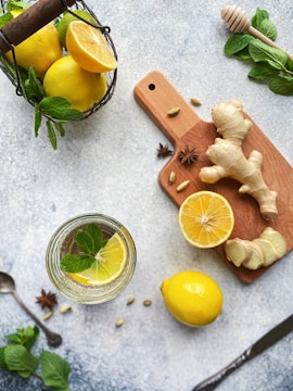 A selection of fresh ingredients including lemons, ginger, and mint is arranged artistically on a textured surface. A glass of water garnished with a lemon slice and mint leaves is placed nearby, while a wooden cutting board holds slices of ginger and a halved lemon. The scene is complemented by a honey dipper and some star anise, creating a refreshing and natural aesthetic.