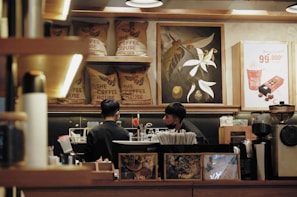 Two people are working behind a counter in a coffee shop. Coffee bean sacks are displayed on a shelf, and there are decorative paintings and a promotional poster in the background. Various coffee-making equipment and supplies are visible around the workspace.