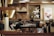 Two people are working behind a counter in a coffee shop. Coffee bean sacks are displayed on a shelf, and there are decorative paintings and a promotional poster in the background. Various coffee-making equipment and supplies are visible around the workspace.
