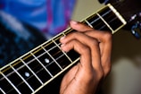 A hand is positioned on the fretboard of a guitar, pressing on the strings to form a chord. The guitar's frets and strings are clearly visible, with the headstock blurred slightly in the background.