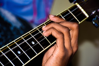 A hand is positioned on the fretboard of a guitar, pressing on the strings to form a chord. The guitar's frets and strings are clearly visible, with the headstock blurred slightly in the background.
