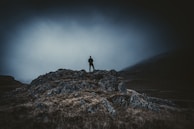 A lone figure cloaked in shadow standing atop a craggy hill under a stormy sky.