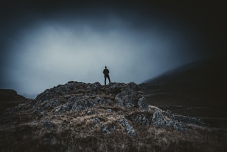 A lone figure cloaked in shadow standing atop a craggy hill under a stormy sky.