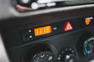 A close-up view of a car dashboard featuring a digital clock displaying the time 11:13. Adjacent to the clock are controls for setting the hours and minutes. Below the clock is a hazard warning light button indicated by a red triangle, and next to it are various climate control dials for air conditioning and heating.