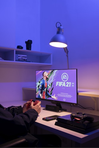 A gaming setup featuring a monitor displaying the FIFA 21 game cover, accompanied by a person holding a game controller. A PlayStation 4 console and another controller are placed on the desk. A desk lamp illuminates the setup with a soft, purple hue. Shelves in the background hold camera lenses and other small items.