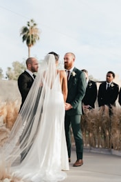 A wedding ceremony is taking place outdoors with a couple standing together holding hands. The bride is wearing a long, white gown with a flowing veil, while the groom is dressed in a green suit with a boutonniere. They are surrounded by pampas grass decorations and accompanied by groomsmen in dark suits. A palm tree and a clear sky serve as the backdrop for the event.
