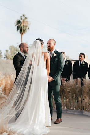 A wedding ceremony is taking place outdoors with a couple standing together holding hands. The bride is wearing a long, white gown with a flowing veil, while the groom is dressed in a green suit with a boutonniere. They are surrounded by pampas grass decorations and accompanied by groomsmen in dark suits. A palm tree and a clear sky serve as the backdrop for the event.