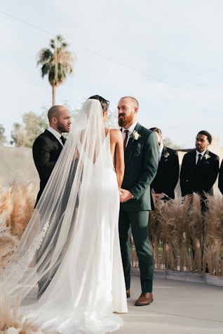 A wedding ceremony is taking place outdoors with a couple standing together holding hands. The bride is wearing a long, white gown with a flowing veil, while the groom is dressed in a green suit with a boutonniere. They are surrounded by pampas grass decorations and accompanied by groomsmen in dark suits. A palm tree and a clear sky serve as the backdrop for the event.