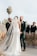 A wedding ceremony is taking place outdoors with a couple standing together holding hands. The bride is wearing a long, white gown with a flowing veil, while the groom is dressed in a green suit with a boutonniere. They are surrounded by pampas grass decorations and accompanied by groomsmen in dark suits. A palm tree and a clear sky serve as the backdrop for the event.