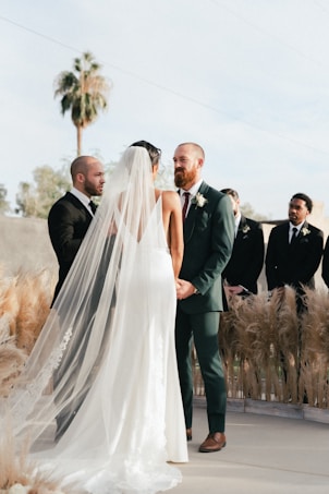 A wedding ceremony is taking place outdoors with a couple standing together holding hands. The bride is wearing a long, white gown with a flowing veil, while the groom is dressed in a green suit with a boutonniere. They are surrounded by pampas grass decorations and accompanied by groomsmen in dark suits. A palm tree and a clear sky serve as the backdrop for the event.