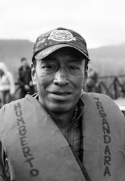 A middle-aged person is wearing a cap and a life jacket with text on it. The background is blurred, showing some people and a natural setting.