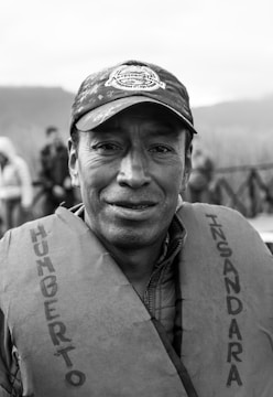A middle-aged person is wearing a cap and a life jacket with text on it. The background is blurred, showing some people and a natural setting.