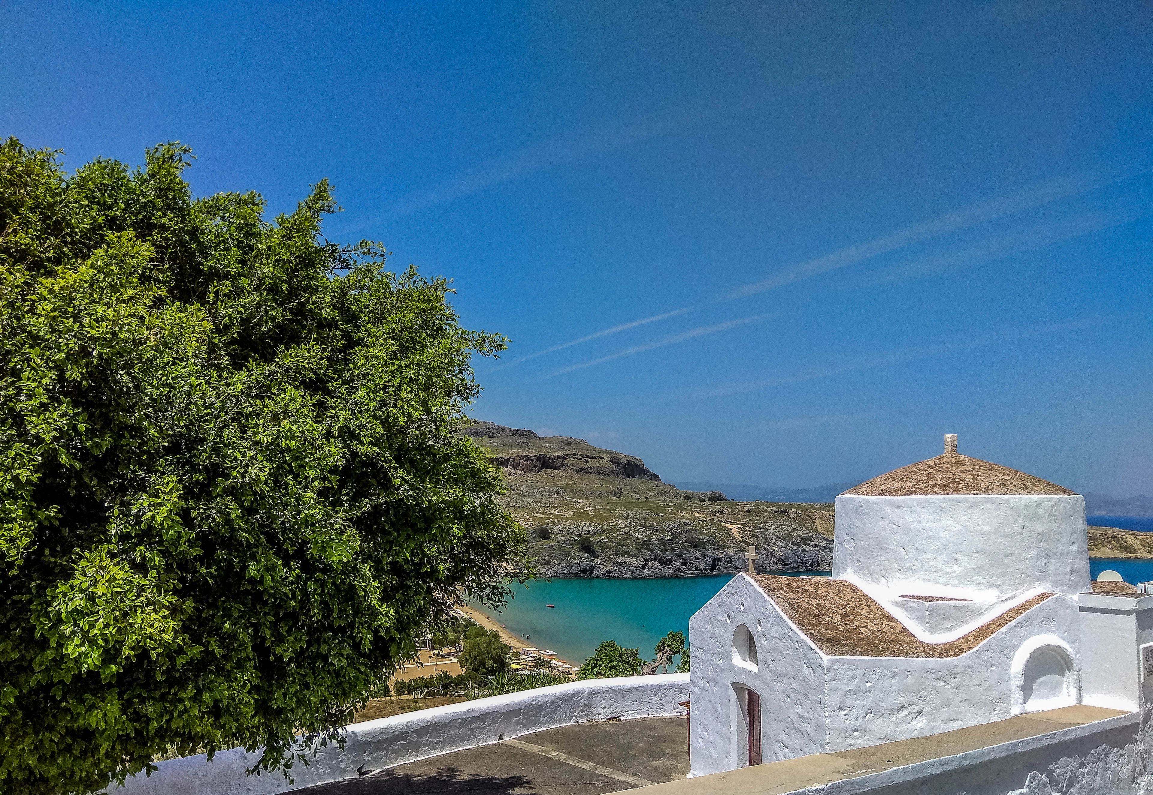 A whitewashed chapel with a domed roof overlooks a turquoise bay in Lindos, framed by a large tree.