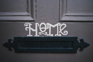 The word 'HOME' is painted in a decorative font on a dark wooden door above a black metal mail slot.