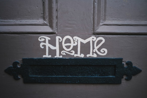 The word 'HOME' is painted in a decorative font on a dark wooden door above a black metal mail slot.
