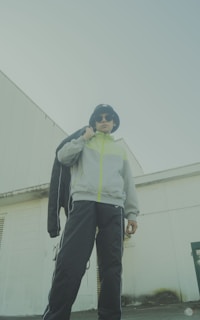 A person wearing a bucket hat, sunglasses, and a light grey jacket with neon accents stands confidently. They are also wearing dark track pants and holding a black jacket over their shoulder. The background includes a white industrial building with geometric lines, lending an urban feel to the scene.