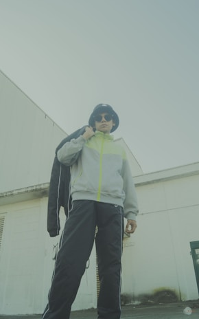 A person wearing a bucket hat, sunglasses, and a light grey jacket with neon accents stands confidently. They are also wearing dark track pants and holding a black jacket over their shoulder. The background includes a white industrial building with geometric lines, lending an urban feel to the scene.