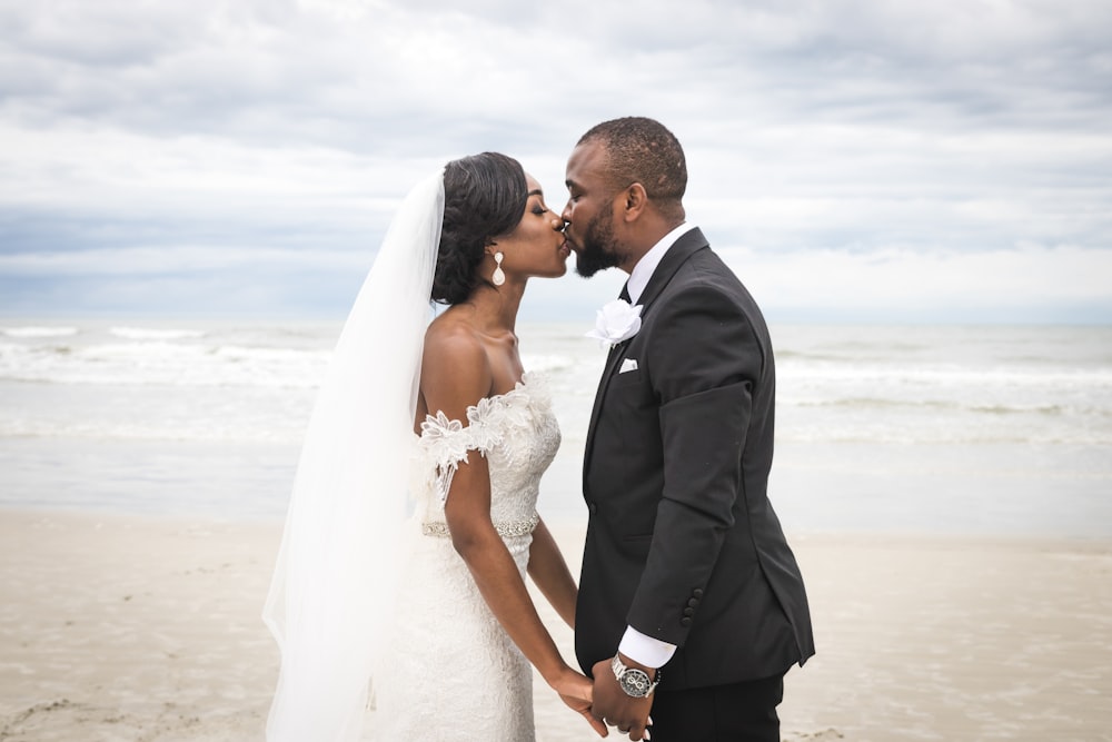 uomo in abito nero che bacia la donna in abito da sposa bianco sulla spiaggia durante il giorno