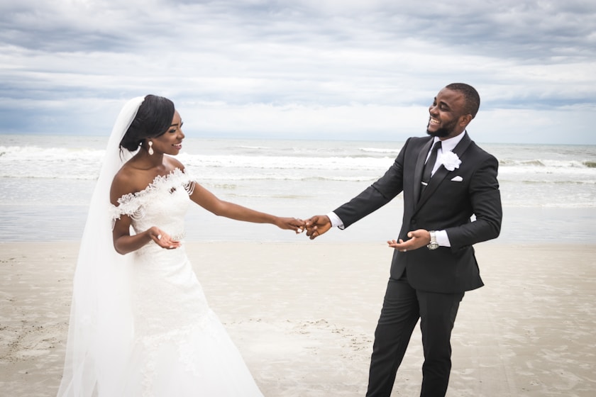 man in black suit holding hands with woman in white wedding dress on beach during daytime