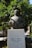 A bronze statue of a man holding a scroll stands in front of lush green trees with visible oranges. The statue is mounted on a white marble pedestal with inscriptions in Greek, indicating dates and a name. The background features more greenery and a partial view of an urban setting.