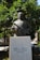 A bronze statue of a man holding a scroll stands in front of lush green trees with visible oranges. The statue is mounted on a white marble pedestal with inscriptions in Greek, indicating dates and a name. The background features more greenery and a partial view of an urban setting.