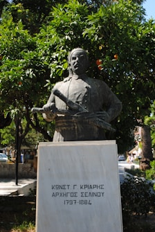 A bronze statue of a man holding a scroll stands in front of lush green trees with visible oranges. The statue is mounted on a white marble pedestal with inscriptions in Greek, indicating dates and a name. The background features more greenery and a partial view of an urban setting.