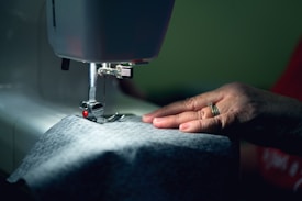 A hand gently holds fabric as it feeds through a sewing machine. The lighting is soft, focusing on the detailed stitching and the textile.